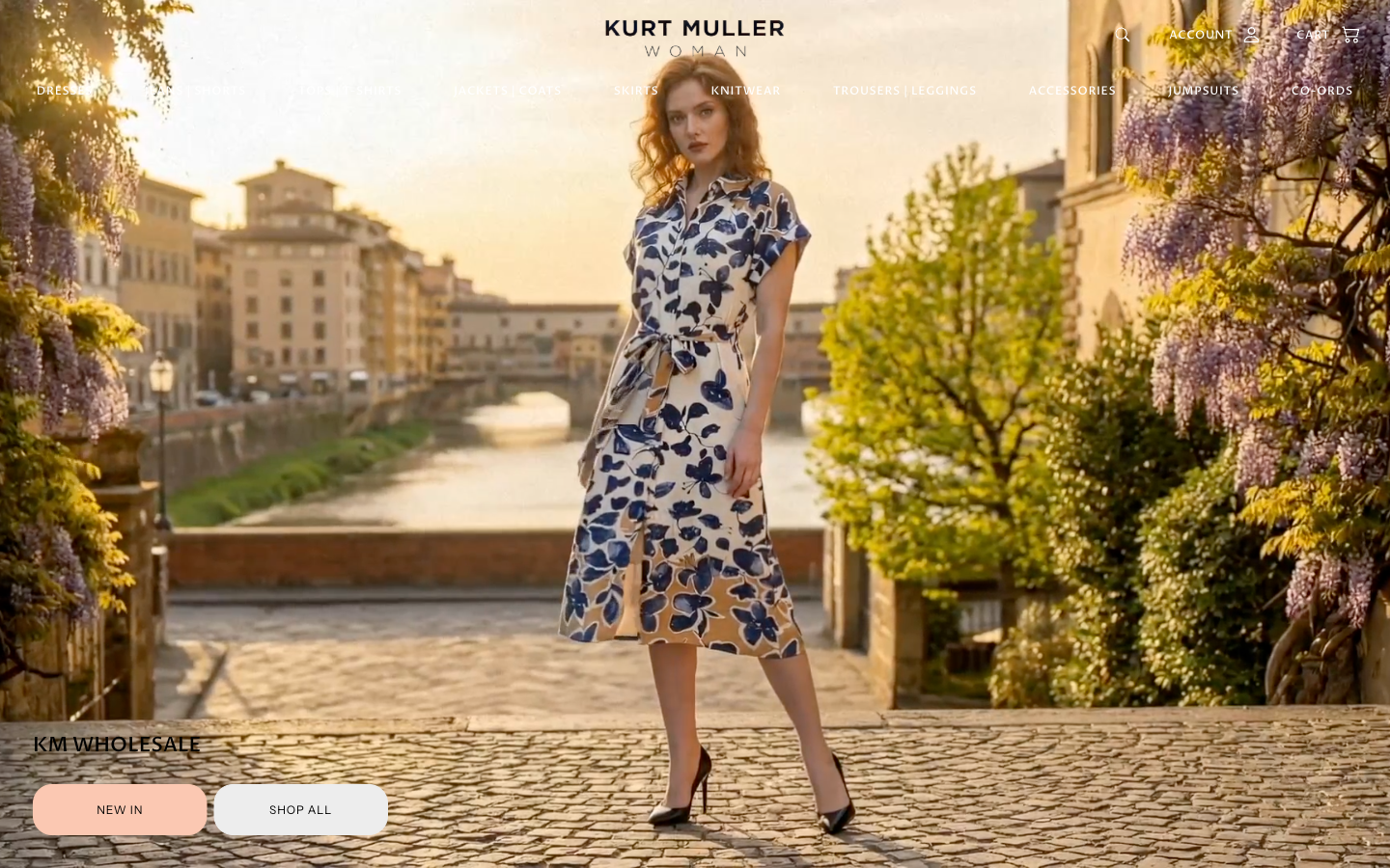 Kurt Muller Wholesale homepage — full-bleed editorial photograph of a model in a navy floral midi dress on a Florence cobblestone street at golden hour, KM WHOLESALE wordmark in the lower-left, cream and shop CTAs in the corner, full womenswear category navigation across the top.
